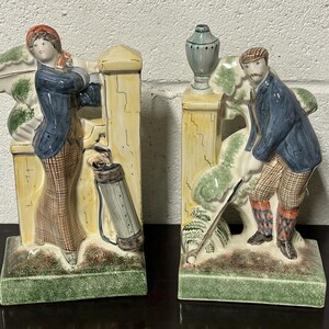 May include: A pair of ceramic bookends depicting a golfer and a woman holding a golf bag. The bookends are painted in a colourful style with green, yellow, blue, and red accents. The bookends are shaped like a fence with a leafy design.