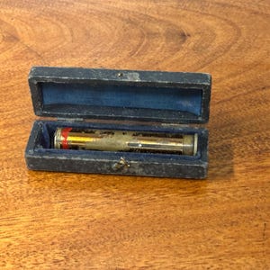 May include: An antique, cylindrical instrument with red and gold accents, displayed in an open, dark blue velvet-lined case. The case is rectangular with a small clasp. The instrument appears to be made of metal and glass.