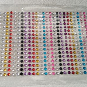 May include: A sheet of self-adhesive gems in various colours, including red, gold, silver, black, purple, pink, and blue. The gems are arranged in rows on a clear plastic sheet.