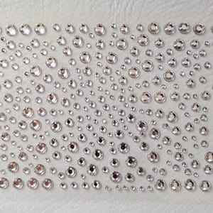 May include: A sheet of clear, round, adhesive rhinestones of various sizes. The rhinestones are arranged in a random pattern on a white background.