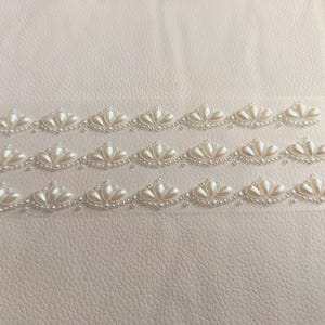 May include: Close-up of rows of off-white, pearl-like embellishments in a fan design. Each fan features a central cluster of larger pearls, encircled by smaller pearls. The background is a light-coloured, textured surface, possibly leather or a similar material.