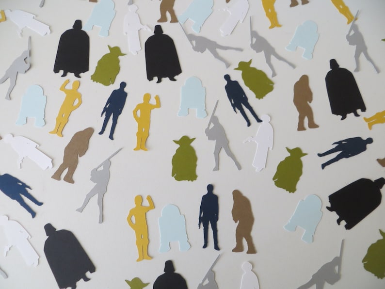 May include: A collection of Star Wars character silhouettes cut from paper in various colors, including black, white, gray, brown, yellow, green, and blue. The characters include Darth Vader, Yoda, Chewbacca, R2-D2, and Luke Skywalker.