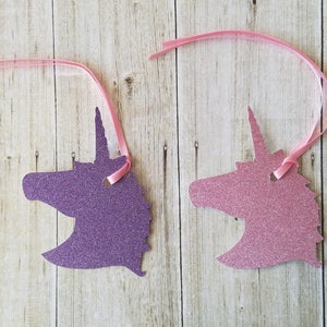 K&ouml;nnte beinhalten: Zwei ausgeschnittene Einhornk&ouml;pfe, einer lila und einer rosa, mit Glitzer-Finish. Jeder Ausschnitt hat ein Loch oben und ist an einem rosa Band befestigt.
