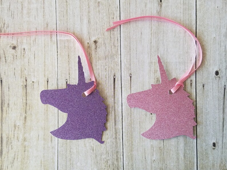 K&ouml;nnte beinhalten: Zwei ausgeschnittene Einhornk&ouml;pfe, einer lila und einer rosa, mit Glitzer-Finish. Jeder Ausschnitt hat ein Loch oben und ist an einem rosa Band befestigt.