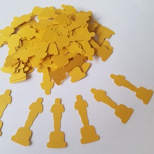May include: A pile of gold-colored paper confetti shaped like award statues. There are also several larger award statue confetti pieces scattered around the pile.