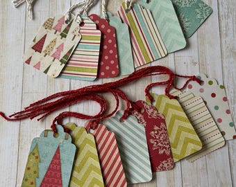 May include: A collection of decorative gift tags with various festive patterns, including Christmas trees, stripes, polka dots, and snowflakes. Each tag is connected to a string, with some using white twine and others using red twine. The tags are arranged on a light wood surface.