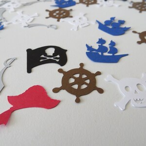May include: Assortment of pirate-themed paper cutouts in various colors, including a red pirate hat, a black pirate flag with a skull and crossbones, a brown ship's wheel, a white skull and crossbones, and blue ships.