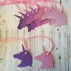 K&ouml;nnte beinhalten: Ein Satz von acht rosa und violetten Glitzer-Einhornkopf-Ausschnitten mit rosa Schleife. Die Einhornk&ouml;pfe sind in verschiedenen Rosat&ouml;nen und Violett.