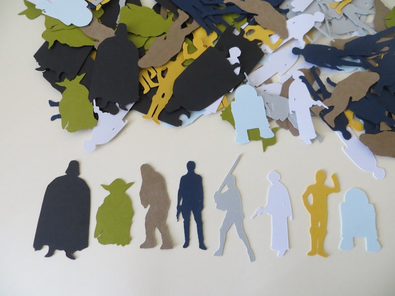 May include: A collection of Star Wars character paper cutouts in various colors, including black, green, brown, blue, gray, white, and gold. The cutouts are arranged in a pile and on a white surface.