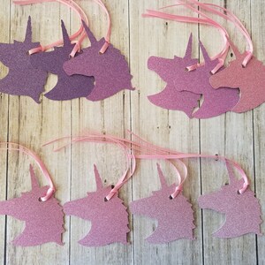 K&ouml;nnte beinhalten: Ein Set mit zehn Geschenkanh&auml;ngern in Form von Einh&ouml;rnern mit pinkfarbenem und violettem Glitzer. Die Anh&auml;nger sind an einem rosa Satinband befestigt.