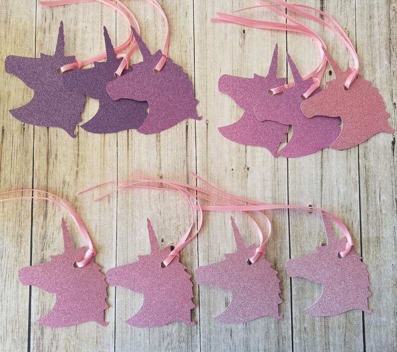 K&ouml;nnte beinhalten: Ein Set mit zehn Geschenkanh&auml;ngern in Form von Einh&ouml;rnern mit pinkfarbenem und violettem Glitzer. Die Anh&auml;nger sind an einem rosa Satinband befestigt.