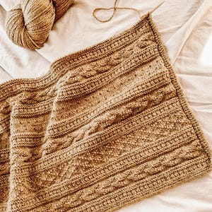 May include: A brown knitted blanket with a cable knit pattern, with two balls of brown yarn beside it. A wooden knitting needle is inserted into the blanket, showing the start of the project. The blanket is on a cream-colored surface.