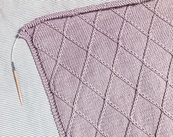 May include: A close-up of a light purple knitted blanket with a diamond pattern. The blanket is being worked on with a wooden knitting needle.