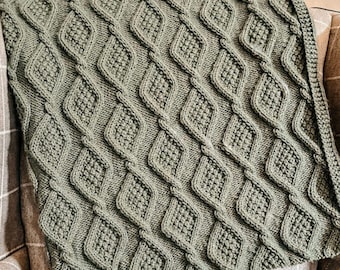 Puede incluir: Una manta de punto verde salvia con un patrón de diamantes, colocada sobre una silla. La textura de la manta es visible, con formas de diamantes en relieve y un borde de punto trenzado. Un elemento de decoración hogareña acogedor.