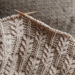 May include: Close-up of a beige knitted fabric with a textured leaf pattern. Two wooden knitting needles are inserted into the top row of stitches. The knitted item is in progress, showing the detailed stitch work.