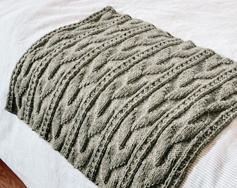 Puede incluir: Una manta de punto color verde salvia que presenta un patrón repetitivo de trenzas verticales con columnas trenzadas texturizadas y pequeños detalles de ojales en toda la tela, creando un diseño de superficie denso y uniforme en toda la pieza rectangular.