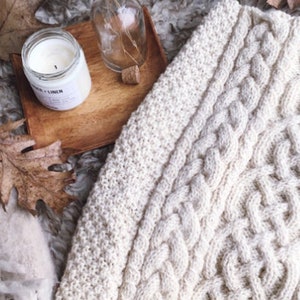 May include: Cream-colored cable knit blanket with a fringed edge. A wooden tray holds a candle, a small glass bottle with dried stems, and a cork. Autumn leaves and a soft, textured surface complete the cozy scene.