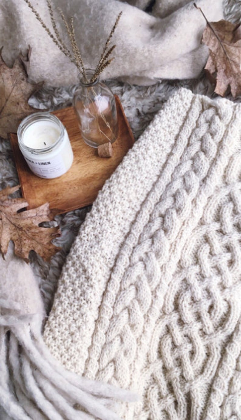 May include: Cream-colored cable knit blanket with a fringed edge. A wooden tray holds a candle, a small glass bottle with dried stems, and a cork. Autumn leaves and a soft, textured surface complete the cozy scene.