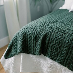 May include: A dark green cable-knit blanket draped over a bed. The blanket has a textured pattern and is paired with a white bed skirt with a subtle, repeating design. The scene is softly lit, suggesting a cozy bedroom setting.