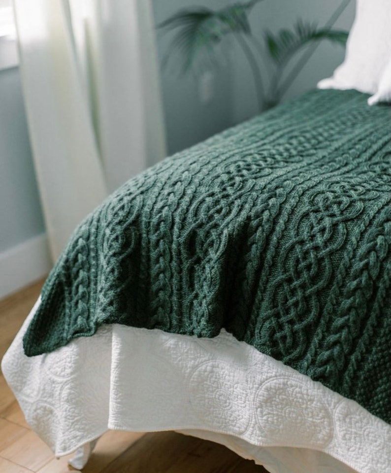 May include: A dark green cable-knit blanket draped over a bed. The blanket has a textured pattern and is paired with a white bed skirt with a subtle, repeating design. The scene is softly lit, suggesting a cozy bedroom setting.