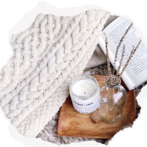 May include: A cozy, cream-colored cable-knit throw blanket is draped over a neutral surface. A wooden tray holds a lit candle labeled "Lavender + Linen", a small glass vase with dried stems, and a book.