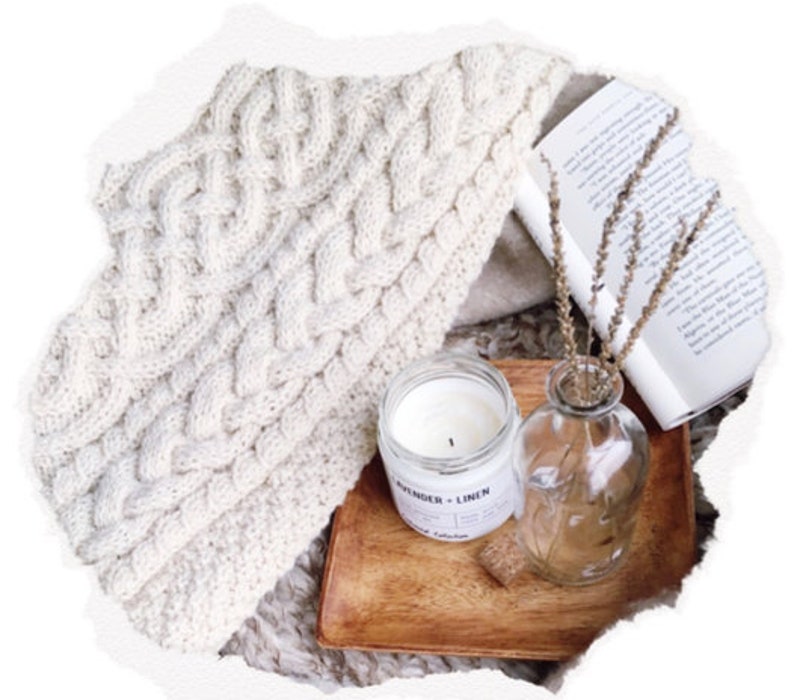 May include: A cozy, cream-colored cable-knit throw blanket is draped over a neutral surface. A wooden tray holds a lit candle labeled "Lavender + Linen", a small glass vase with dried stems, and a book.