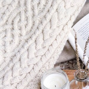 May include: Cream-colored cable knit blanket with a thick, textured weave. A lit candle, a sprig of dried lavender, and an open book are arranged on a wooden tray, creating a cozy scene. The blanket is folded, showing its intricate pattern.