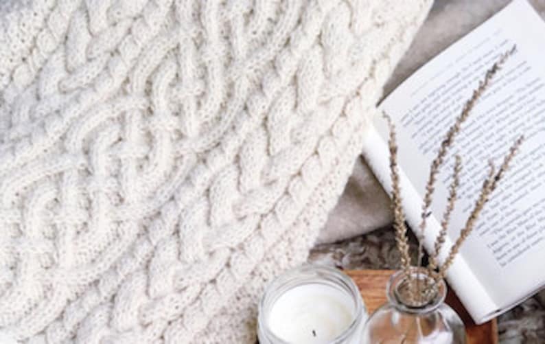 May include: Cream-colored cable knit blanket with a thick, textured weave. A lit candle, a sprig of dried lavender, and an open book are arranged on a wooden tray, creating a cozy scene. The blanket is folded, showing its intricate pattern.