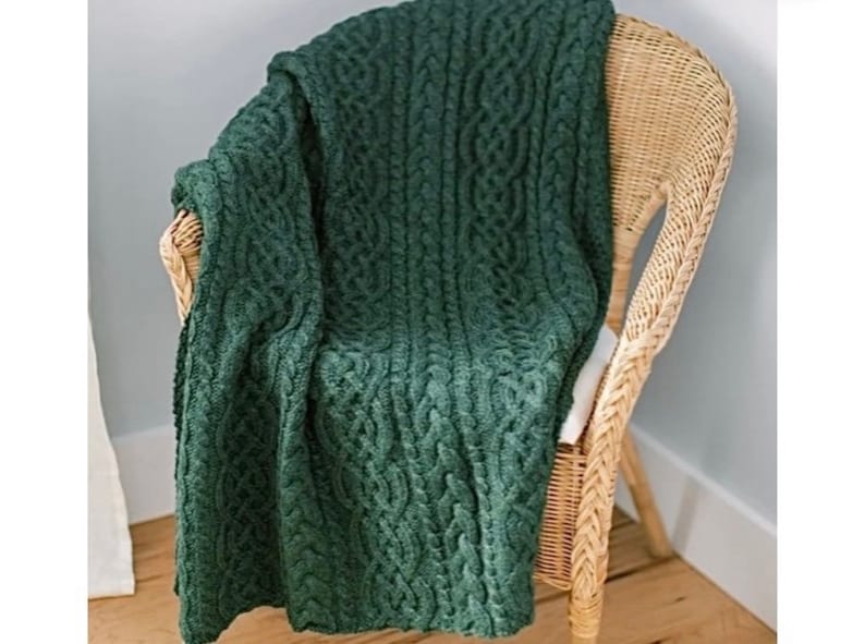 May include: A dark forest green knitted throw blanket featuring a prominent vertical cable knit pattern. The blanket is draped over a light-colored woven wicker chair, showcasing the textured, chunky yarn construction and deep green color.