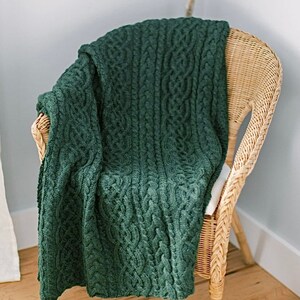 May include: A dark green cable-knit throw blanket draped over a wicker chair. The blanket features a detailed woven pattern and is made of a soft, textured material. The chair has a natural, light brown finish.