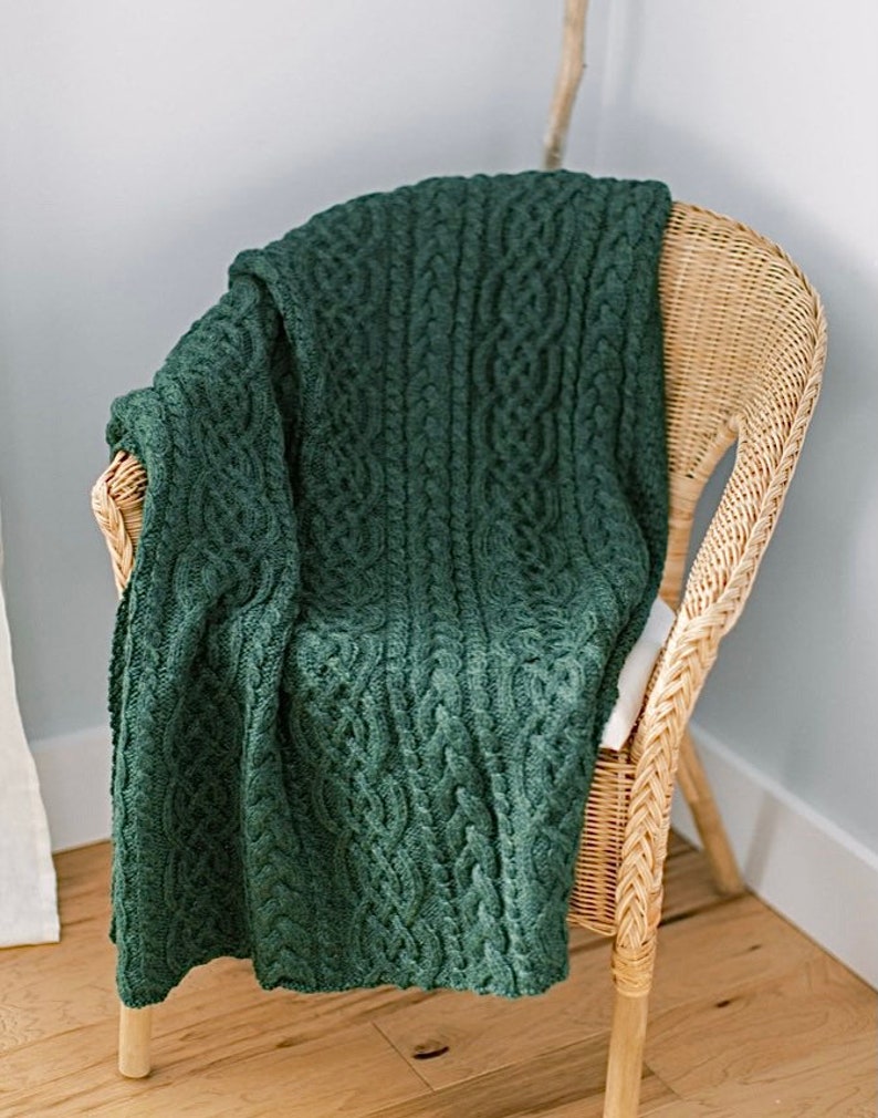 May include: A dark green cable-knit throw blanket draped over a wicker chair. The blanket features a detailed woven pattern and is made of a soft, textured material. The chair has a natural, light brown finish.