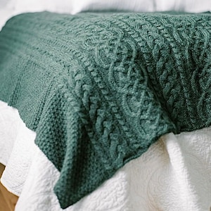 May include: A dark teal knitted blanket with a cable knit design. The blanket is draped over a white bedspread, creating a cozy and inviting aesthetic. The intricate pattern adds texture and visual interest to the bedding.