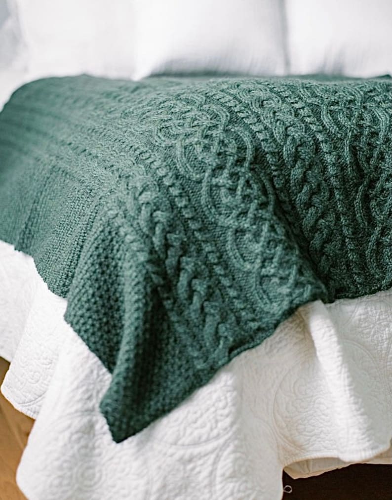 May include: A dark teal knitted blanket with a cable knit design. The blanket is draped over a white bedspread, creating a cozy and inviting aesthetic. The intricate pattern adds texture and visual interest to the bedding.