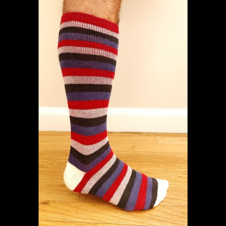 May include: A knee-high sock with horizontal stripes in red, purple, black, gray, and white. The sock is worn on a foot and leg, with the stripes wrapping around the leg and foot. The toe of the sock is white.