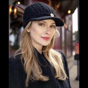 May include: A black knit beanie with a brim and a gold button on the top. The beanie is worn by a woman with long blonde hair.