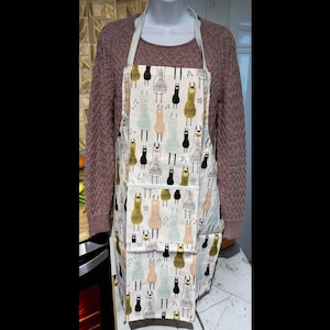 Puede incluir: Un delantal blanco con un patrón repetido de llamas negras, doradas y celeste. El delantal tiene dos bolsillos y correas celestes. El delantal se muestra en un maniquí.