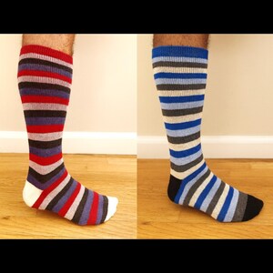 May include: Two knee-high socks, each with horizontal stripes. One sock features red, purple, gray, and white stripes. The other sock has blue, gray, and white stripes. Both socks are on a wooden floor.