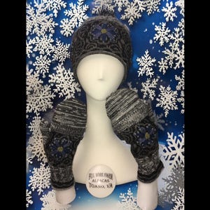 May include: A black and white knit hat with a blue flower design and matching mittens. The hat has a pom pom on top. The mittens have a blue flower design on the back. The hat and mittens are displayed on a white mannequin against a blue background with white snowflakes.