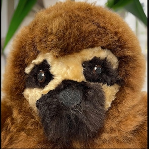 May include: Close-up of a brown and tan plush sloth toy with black eyes and a black nose. The toy has a soft, furry texture.