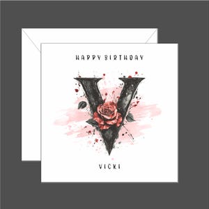 May include: A white card with the text "Happy Birthday" and the letter "V" in black with a red rose and pink watercolour splatter. The name "Vicki" is written below the letter.