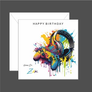 May include: A birthday card with a colourful design featuring a video game controller and headphones with colourful paint splatters. The text "Happy Birthday" is at the top of the card and the text "Game On Zak" is at the bottom of the card.