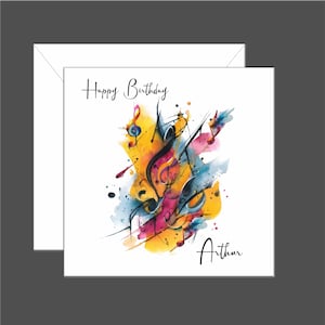 May include: A white birthday card with the words "Happy Birthday" and "Arthur" in black script. The card features a colourful watercolour design of musical notes in yellow, orange, pink, and blue.
