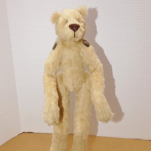 May include: A beige plush teddy bear with brown eyes and a brown nose. The bear has a long, slender body and long arms and legs. The bear is standing on a brown surface.