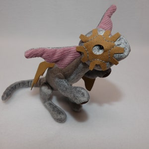May include: A gray plush creature with pink corduroy wings and a brown gear-shaped eye mask. The handmade toy has brown leather-like accents and is posed on a white surface. The stuffed animal has a whimsical, fantasy theme.