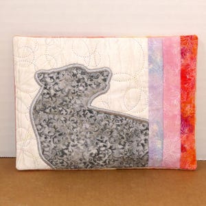 May include: A rectangular quilted item with a bear design. The bear is grey with a floral pattern, set against a cream background with stitched circles. The right side has vertical stripes of pink, purple, and orange fabrics.