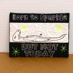 May include: A rectangular, quilted wall hanging with the text "Born to Sparkle" and "But Not Today". It features a white dinosaur illustration on a white background with star and zigzag patterns, and neon green accents.