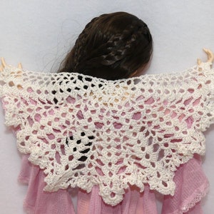 Pode incluir: Xale branco de crochê com um delicado padrão de renda. O xale está drapeado sobre uma boneca que veste um vestido rosa.