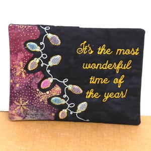 May include: A rectangular, black fabric item with the embroidered text "It's the most wonderful time of the year!". The left side features a purple and gold snowflake pattern, with a string of colorful lights.
