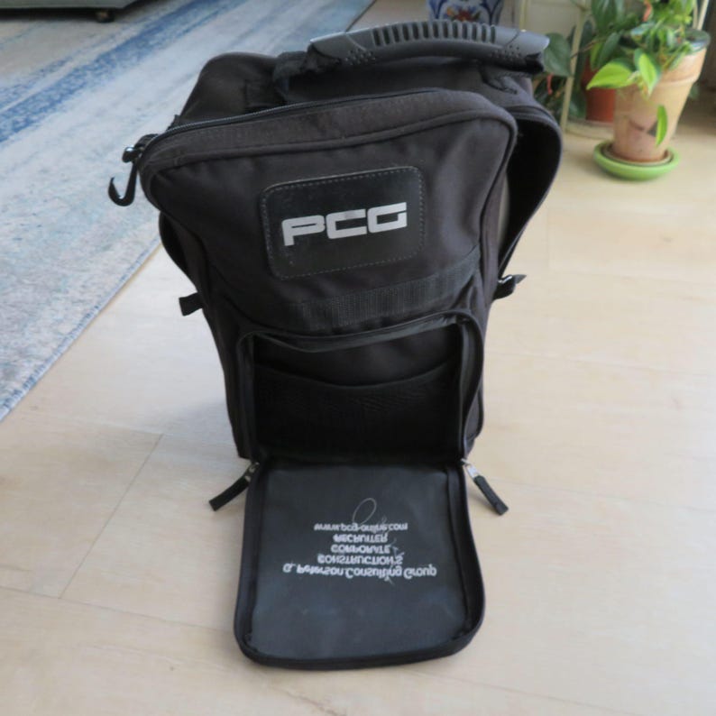 May include: Black backpack with the letters "PCG" in a white rectangle on the front. The bag has multiple compartments, a handle, and a zippered opening. The bottom compartment is open, revealing text.