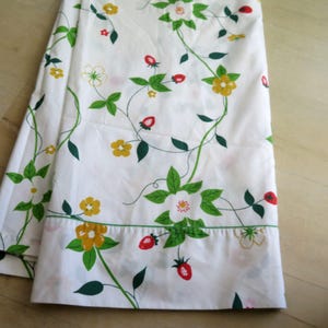 Puede incluir: Tela blanca con un patr&oacute;n repetido de vides verdes, hojas, flores amarillas, flores blancas y fresas rojas. La tela est&aacute; doblada, mostrando el patr&oacute;n en detalle. El dise&ntilde;o es un patr&oacute;n floral y de frutas.